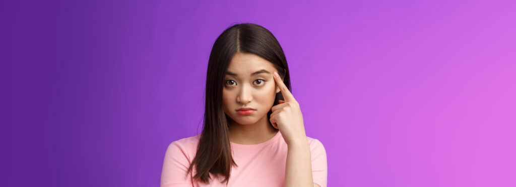 Shocked asian girl scold friend unacceptable behaviour, pointing finger temple look confused and upset stare camera displeased weird actions, asking are you stupid, stand purple background.