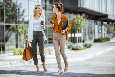 Full body portrait of a two young businesswomen walking with coffee cups near the modern office building outdoors