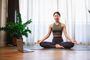 Asian woman practicing yoga from yoga online course on a video conference on a laptop at home. Healthy lifestyle - technology at home. New normal lifestyle, Resilienz Training, Resilienz Übung