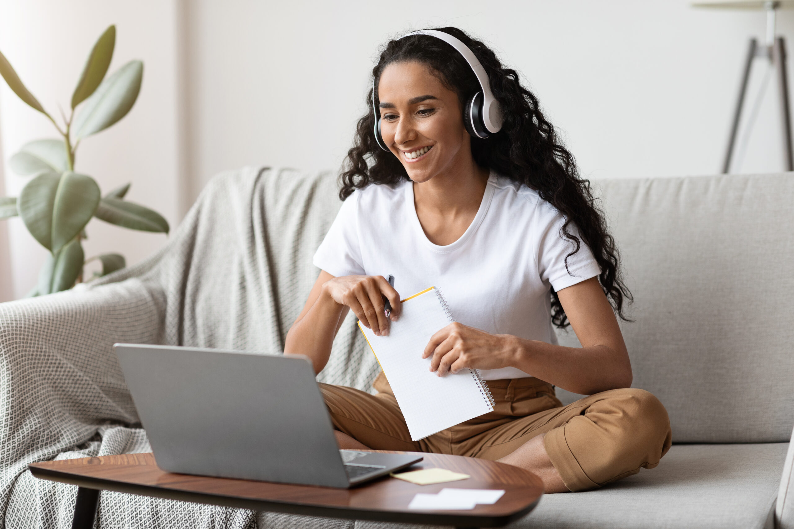 Cheerful pretty brunette lady attending online language course from home, sitting on sofa, using laptop and wireless headset, taking notes and smiling, enjoying online studying, copy space
