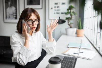 Angry young businesswoman fighting with assistant or business partner, talking on cellphone and gesturing, having difficulties with business, sitting in coworking space